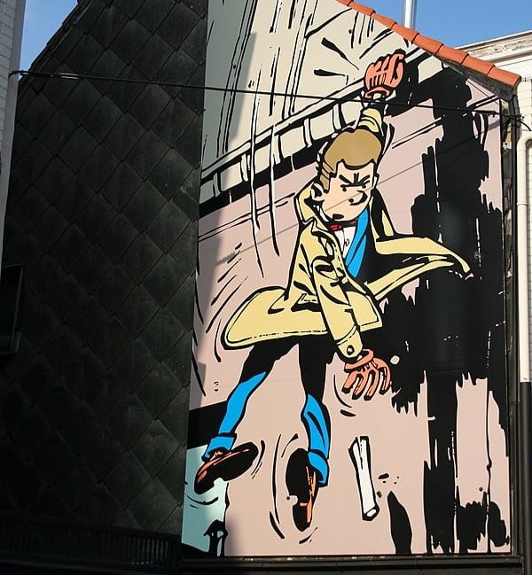A mural in Brussels, Belgium depicting a cartoonish man holding on to the edge of the roof with his left hand while dropping a piece of paper.