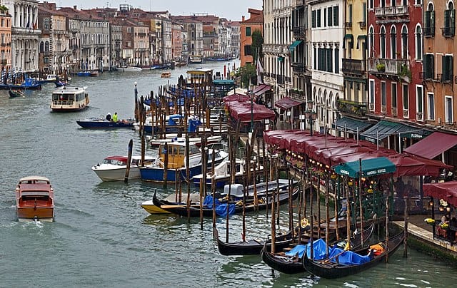 Gondolas parked on the side of the canal just in front of restaurants while other boats pass by.