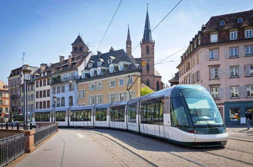 A tram in Strasbourg in the middle of the street running on a track with overhead electric wires with buildings in the background