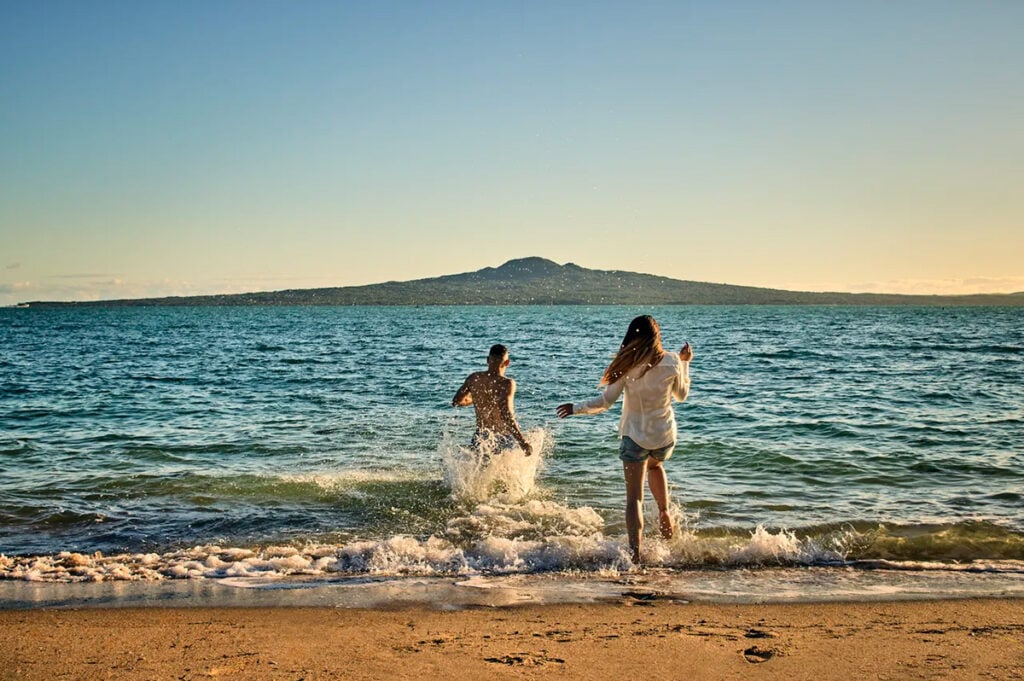 A couple splashing in Mission Bay Beach with Rangitoto Island in front of them