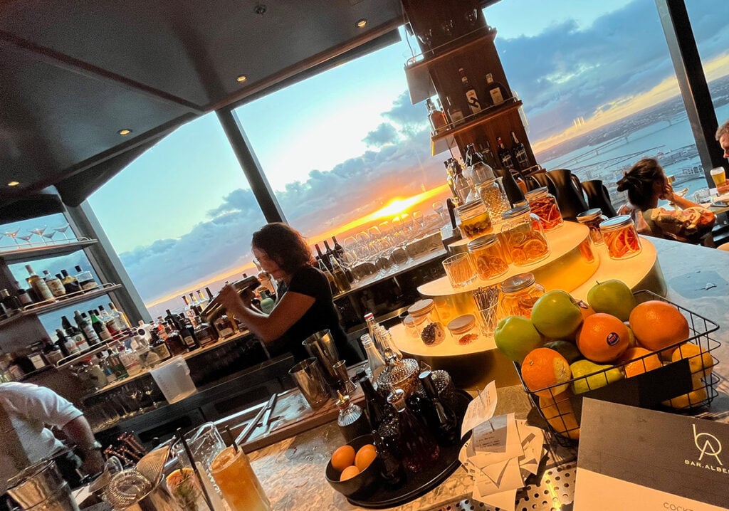 A mixologist shakes a cockail at Bar Albert rooftop as the sun sets over the Auckland Harbour Bridge
